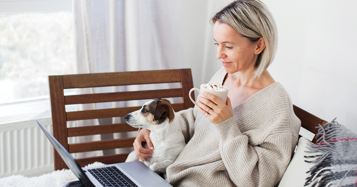 Woman with dog on her lap using laptop while holding a mug of cocoa