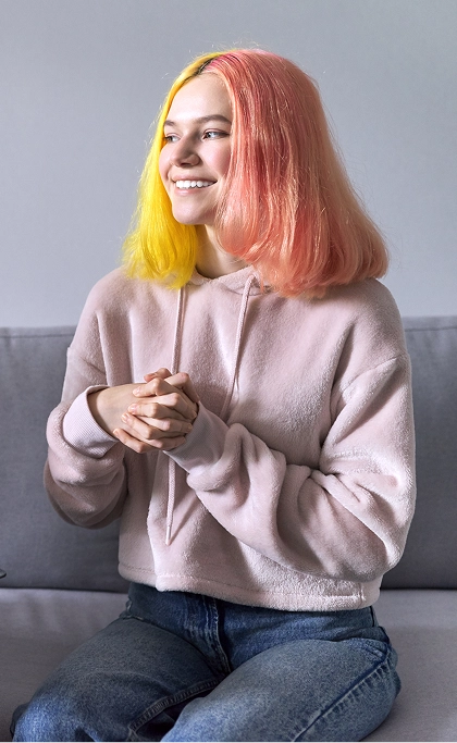 Smiling young person with pink and yellow hair sitting on a couch