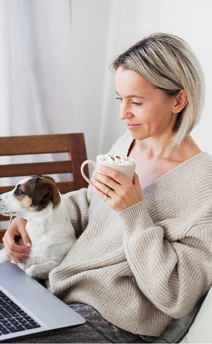Woman holding mug of cocoa with dog on her lap while working on laptop