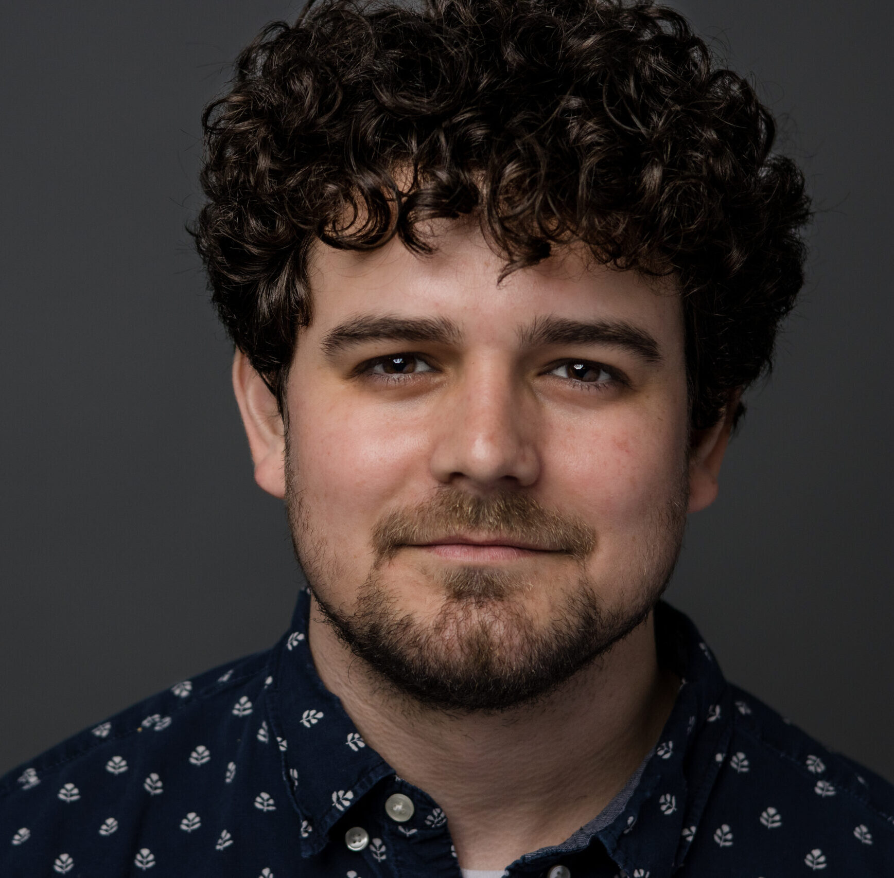 Professional headshot of a man with curly hair and beard against gray background