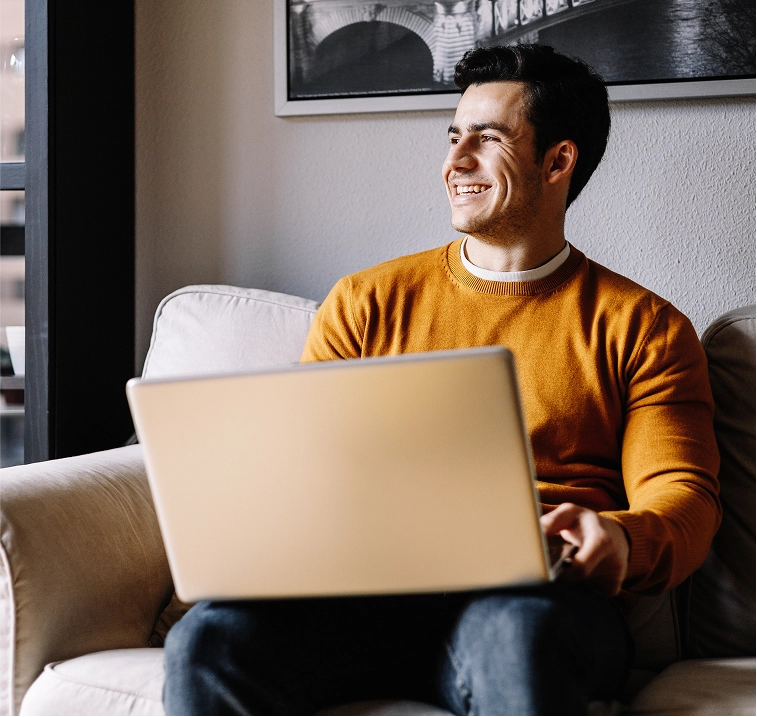 Smiling man in an orange sweater sitting on a couch with a laptop, looking to the side with a relaxed expression