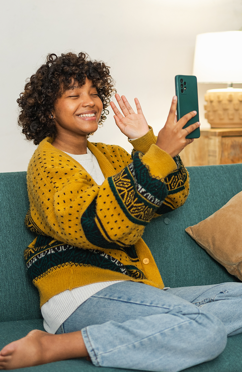 Smiling woman sitting on couch waving during video call on smartphone